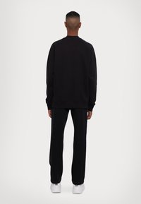 Schwarzer Pullover mit Rundhalsausschnitt, gerippten Bündchen und Saum. Kombiniert mit weiten schwarzen Hosen und weißen Turnschuhen, von hinten betrachtet.