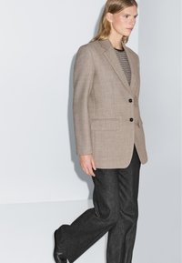 Beige op maat gemaakte blazer van gestructureerde stof met twee zwarte knopen, gecombineerd met donkergrijze wijde broek en een gestreepte top eronder.