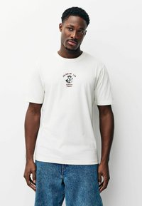 Fehér pamut rövid ujjú póló, középen piros és fekete színű alakzat és szöveg látható. Kék farmernadrággal viselve.