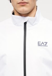 Το κάτω μέρος του προσώπου και ο λαιμός ενός άνδρα που φοράει λευκό μπουφάν με φερμουάρ και το λογότυπο EA7 Emporio Armani στο στήθος.