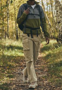 Pullover in pile verde scuro e grigio, pantaloni cargo beige con tasche e scarpe da trekking chiare. Sentiero di camminata circondato da alberi.