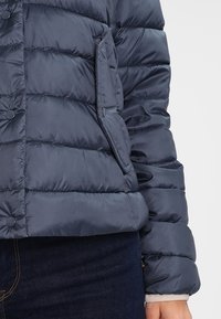 Giacca invernale trapuntata blu navy con bottoni a pressione e tasca con patta indossata su un torso e un braccio di una persona, abbinata a jeans in denim scuri.
