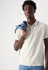 Beige pólóing navy kiegészítőkkel, három gombos kivágással és diszkrét logóval. A modell a vállán egy farmerdzsekit tart.