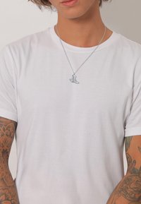 Sølv vedhængs halskæde med et lille detaljeret design af en støvle, båret over en simpel hvid T-shirt, med synlige arm tatoveringer.