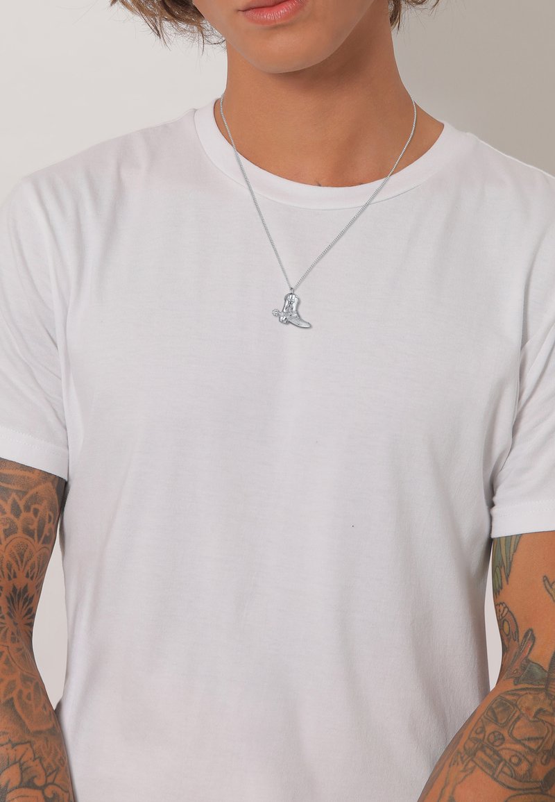 Sølv vedhængs halskæde med et lille detaljeret design af en støvle, båret over en simpel hvid T-shirt, med synlige arm tatoveringer.