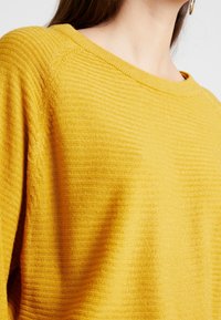 Żółty sweter o fakturze z szerokim dekoltem i prążkowanym wzorem; miękki materiał, luźny krój oraz delikatnie widoczne detale szwów.