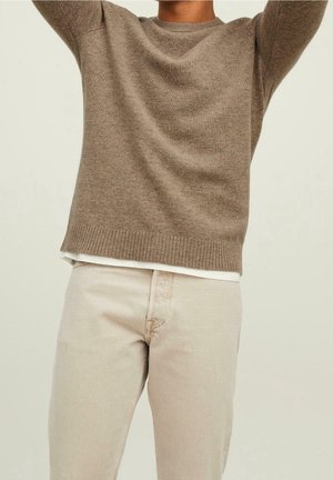 Personne portant un pull en maille marron et un pantalon beige, bras levés, cadrée du cou aux cuisses sur un fond uni.