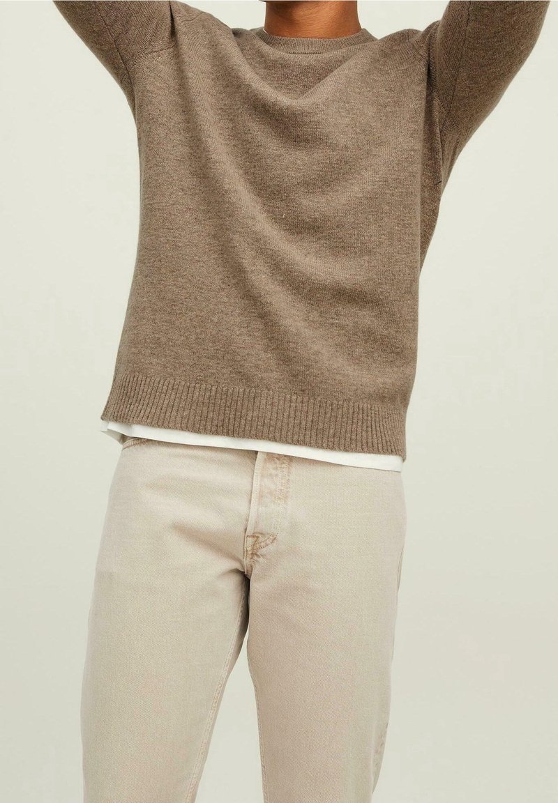 Personne portant un pull en maille marron et un pantalon beige, bras levés, cadrée du cou aux cuisses sur un fond uni.