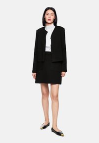 Black textured blazer and matching skirt outfit; features front pockets and a collared white blouse underneath; black flats with gold accents.