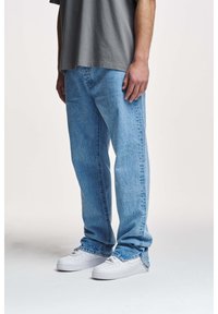 Jeans en denim bleu clair, amples, avec une texture délavée et des ourlets retroussés, associés à des baskets blanches. Design décontracté, coupe droite.