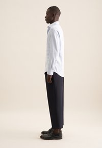 Camicia a maniche lunghe a righe azzurro chiaro abbinata a pantaloni scuri su misura e scarpe nere. La camicia ha un colletto aperto e un orlo curvo.