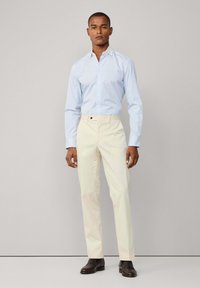 Uomo in piedi contro uno sfondo grigio, che indossa una camicia a righe azzurri chiaro, pantaloni crema e scarpe eleganti marrone scuro.