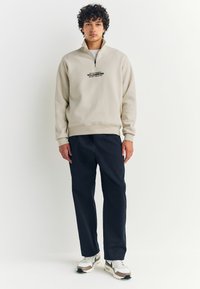 Hellbeiger Sweatshirt mit Reißverschluss und hohem Kragen, schwarzem Text, kombiniert mit dunkelblauen Hosen und weißen Schuhen mit beigen Akzenten.