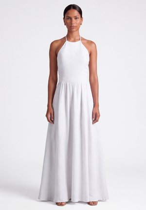 Donna con capelli scuri che indossa un abito lungo bianco senza maniche con collo halter, in piedi davanti a uno sfondo bianco uniforme.