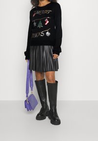 Schwarzer Weihnachts-Sweater mit metallischem Stickerei, schwarzer, plissierter Kunstlederrock, kniehohe schwarze Stiefel und eine lila Handtasche.