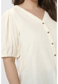 Blouse couleur crème avec un col en V, des manches bouffantes et de petits boutons bruns. Tissu doux et texturé avec une coupe décontractée.