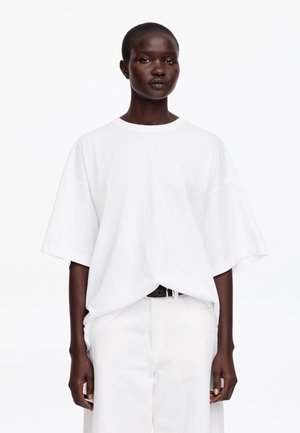 Donkere huidkleurige persoon draagt een oversized eenvoudige witte T-shirt ingestopt in een witte broek met hoge taille en zwarte riem, staand tegen een witte achtergrond.