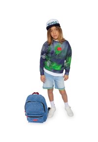 Blauwe denim rugzak met rood logo en groene accenten. Kind in een gestreepte, grafische sweatshirt, lichtblauwe shorts en witte sneakers.