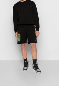 Schwarzer Sweatshirt mit kleinem weißen Logo, kombiniert mit schwarzen Shorts, die ein farbenfrohes Grafikdesign an der Seite aufweisen. Dazu werden gemusterte Sneakers getragen.