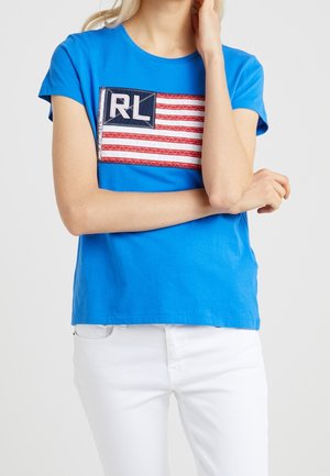 Kvinde iført en blå t-shirt med et syet rødt og hvidt stribet flag og bogstaverne "RL", kombineret med hvide bukser, armene krydsede.