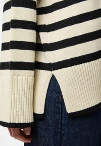 Sweter w paski w kolorze kremowym i czarnym, z ściągaczami i rozcięciami po bokach. Dzianinowa tekstura, luźny krój oraz kontrastowy czarny brzeg przy rękawach.