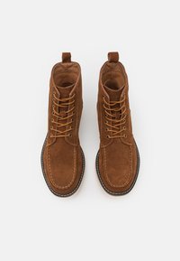 Botas de ante marrón hasta el tobillo con punteras redondeadas, cordones marrones, ojetes dorados y un talón reforzado. Costuras en contraste y una suela duradera.