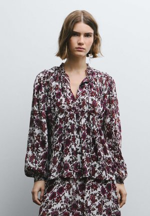 Mujer con cabello corto, que lleva una blusa de manga larga con estampado floral y una falda a juego en tonos blancos, morados y rojos, de pie frente a un fondo gris liso.