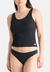 Vrouw in een zwart geribbeld tanktop en zwarte naadloze ondergoed met Adidas-logo's, staand tegen een effen lichte achtergrond.