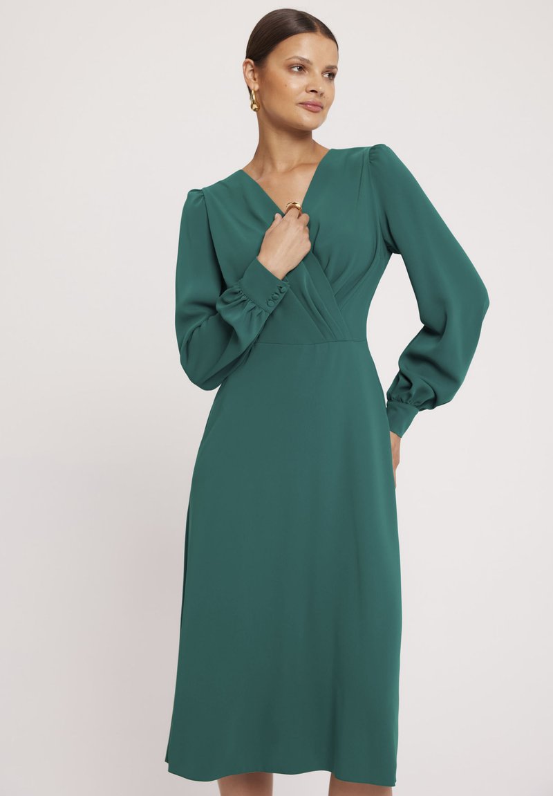 Turkusowa sukienka z długimi, bufiastymi rękawami, dekoltem w kształcie V i plisowanym przodem. Gładka faktura, długość do kolan z delikatną sylwetką w kształcie litery A.