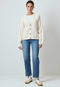 Cardigan beige con scollo a V, apertura frontale con bottoni, due tasche frontali e polsini a coste, abbinato a jeans blu a gamba dritta e sneaker bianche.