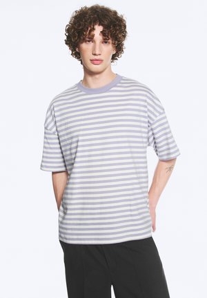 Jeune personne aux cheveux bouclés portant un t-shirt à manches courtes rayé violet clair et blanc et un pantalon noir, debout les mains dans le dos.