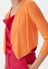 Pomarańczowy dziany kardigan z otwartym przodem, z prążkowaną fakturą i rękawami do łokcia, noszony na różowej satynowej bluzce i różowych spodniach.