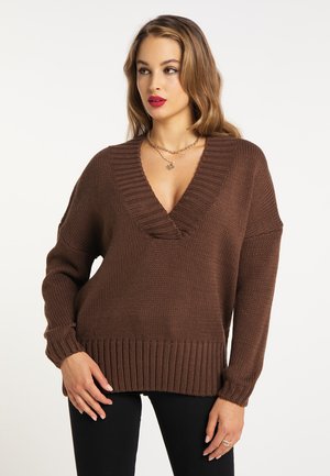 Donna che indossa un maglione marrone a collo a V lavorato a maglia e pantaloni neri, con collane a strati e rossetto rosso, in piedi su uno sfondo bianco.