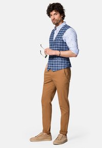 Męska granatowa kamizelka w kratę noszona na jasnoniebieskiej koszuli w wzory, zestawiona z brązowymi chinosami i beżowymi sneakersami. Dodatki obejmują okulary przeciwsłoneczne.