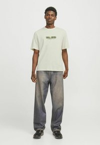 Lys beige bomulds t-shirt med grafisk print i grøn og sort, parret med løstsiddende blå denim jeans og sorte sko.