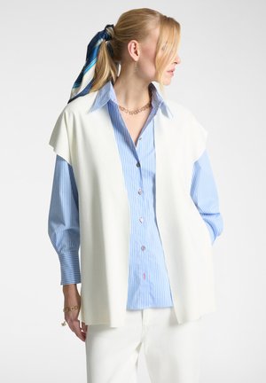 Canottiera bianca senza maniche sopra una camicia blu a righe con bottoni, caratterizzata da una vestibilità sartoriale, colletto e ampie maniche, abbinata a pantaloni bianchi.
