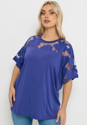 Frau trägt ein locker sitzendes, königsblaues Oberteil mit durchscheinender, floraler Spitze an Schultern und Ärmeln, kombiniert mit hellblauen Jeans, steht vor schlichtem Hintergrund.