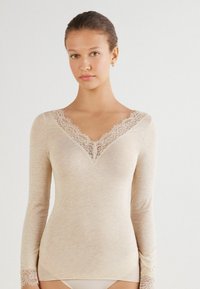 Beige långärmad topp i mjukt tyg, med en spetskantad V-ringning och spetsdetaljer vid ärmslut för extra finess.