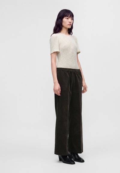 Jeune femme debout portant un haut en maille crème à manches courtes, un pantalon large en velours côtelé vert foncé et des bottines noires à talons.