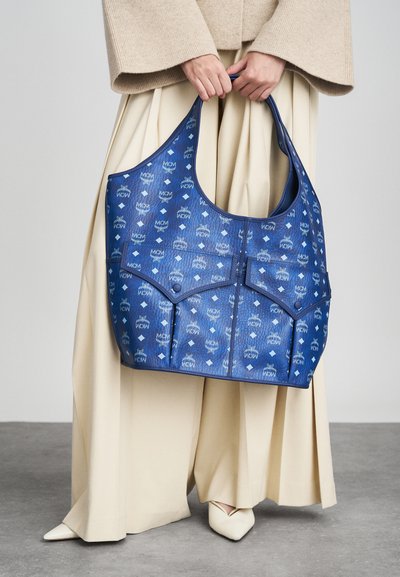 Μπλε τσάντα tote φτιαγμένη από σχεδιασμένο δέρμα με λογότυπα, καμπύλη μορφή, μεγάλο ανοιχτό πάνω μέρος και μπροστινή τσέπη με φερμουάρ.