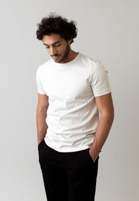 T-shirt bianca in cotone con maniche corte, colletto rotondo e design aderente. Abbinata a pantaloni neri, il modello ha una posa neutra.