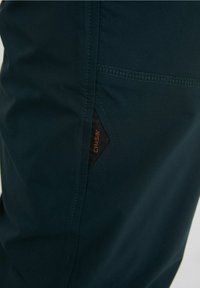 Mörk tealbyxor med en slät yta, blixtlås-fickor på sidan och en svart etikett med orangea bokstäver som varumärkesdetalj.