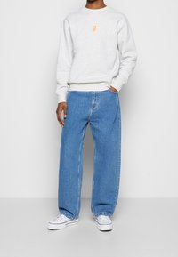 Sudadera gris con logo naranja, combinada con jeans de mezclilla azul de corte holgado. El modelo lleva zapatillas blancas. Diseño simple y casual.