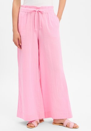 Personne portant un pantalon rose large avec une taille à cordon et des sandales plates roses à bout ouvert, debout devant un fond uni.