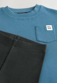 Blauwe geribde longsleeve shirt met zakdetail, gecombineerd met zwarte geribde broek, die een gestructureerde stof en contrasterende kleuren tonen.