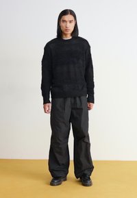 Black knit sweater with textured patterns, paired with loose-fitting black trousers. Footwear consists of black sturdy shoes.