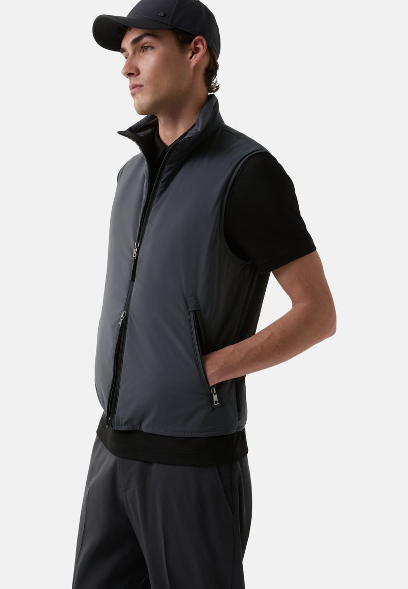 Grau ärmellose Weste aus leichtem Material, mit hohem Kragen, Frontreißverschluss und zwei seitlichen Reißverschlusstaschen. Über einem schwarzen Shirt getragen.