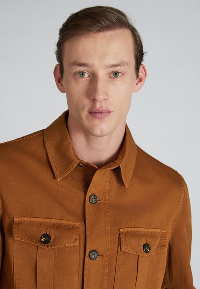 Camisa marrón de botones con dos bolsillos en el pecho, hecha de tela suave, con un cuello clásico y botones negros en la parte delantera.