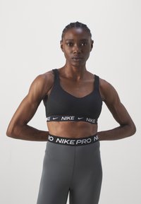Svart sportsbh med V-halsdesign og brede stropper. Har en elastisk bånd med merkevaren "NIKE" i hvitt. Kombinert med grå tettsittende leggings.