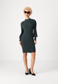 Πλεκτό φόρεμα με ραβδώσεις σε σκούρο πράσινο, με υψηλή λαιμόκοψη, φουσκωτά μανίκια και εφαρμοστή σιλουέτα. Φοριέται με μεγάλα ηλιόλουστα γυαλιά και μαύρες γόβες.